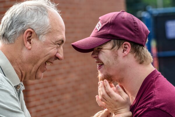Image of an older man smiling at a young man with Down's Syndrome. The young man is smiling back at him. The older man is wearing a grey button up shirt, and then young man has a burgundy tee and a matching hat.